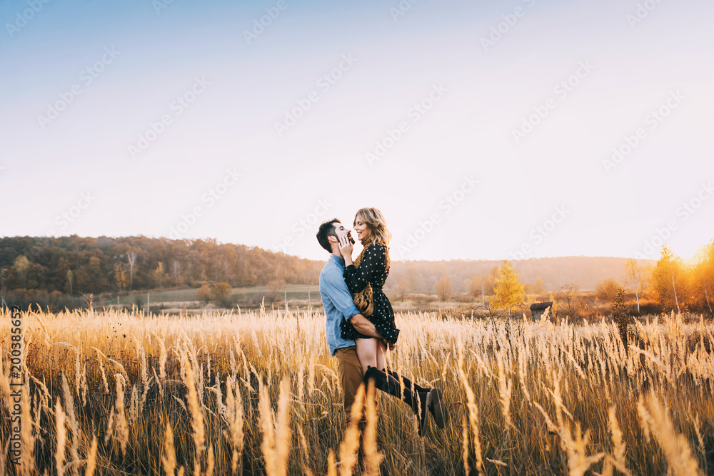 handsome guy with a beard in a blue denim shirt gentle hugs, hand holding and kissing a girl with blond hair in a blue dress and yellow scarf in a field at sunset. stylish couple - obrazy, fototapety, plakaty 