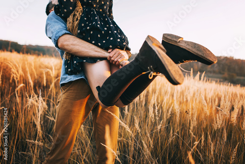 handsome guy with a beard in a blue denim shirt gentle hugs, hand holding and kissing a girl with blond hair in a blue dress and yellow scarf in a field at sunset. stylish couple