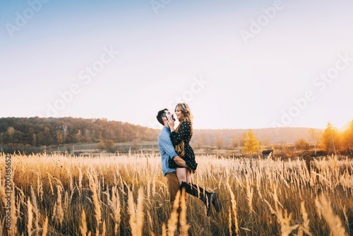 handsome guy with a beard in a blue denim shirt gentle hugs, hand holding and kissing a girl with blond hair in a blue dress and yellow scarf in a field at sunset. stylish couple © Oleksandr