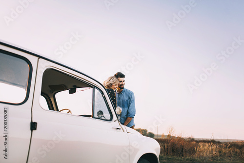 Young beautiful couple hugging each other, sitting on a small white car in beautiful evening light. Stylish guy with a beard and blond girl laughing