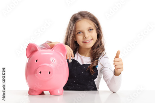 Little girl with a piggybank sitting at a table and making a thumb up gesture