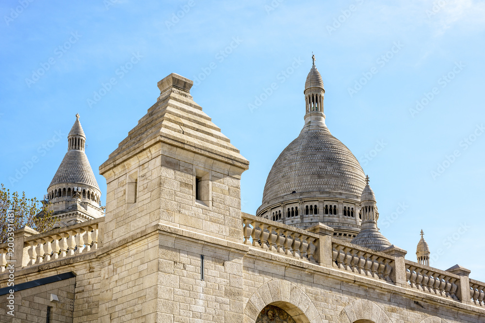 View of the dome and steeple of the Basilica of the Sacred Heart of Paris with the outer stone wall and a turret of the great water reservoir of Montmartre in the foreground under a clear blue sky.