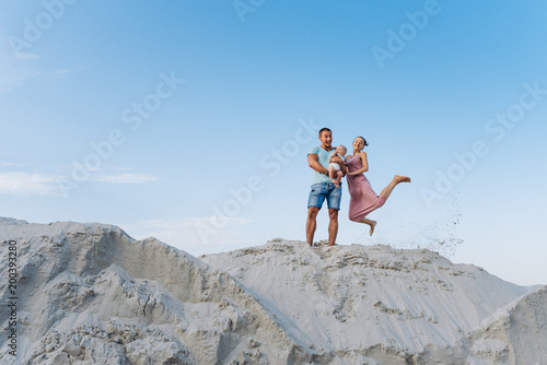 Happy emotional family: mother, father and little baby boy having fun outside on sand on blue sky background