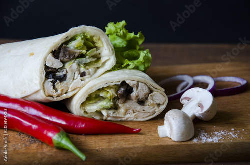 Mexican Food Burrito with chicken, mushrooms, salad and sauces with a red chili peppers and cut mushrooms onion on wooden platform with blue paper background. Tasty food with a vegetables and meat.