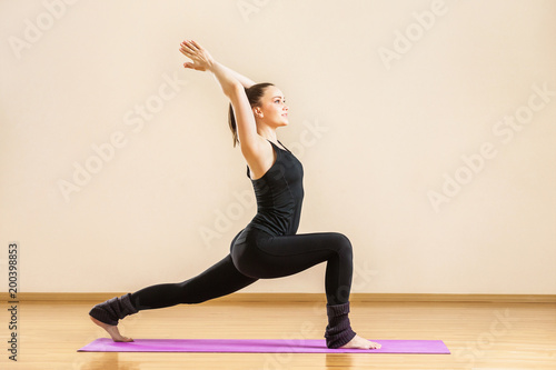 Young woman is practising virabhadrasana known as warrior yoga pose at gym background.