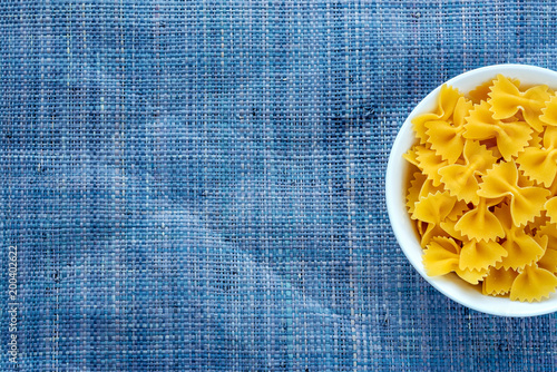 farfalle macaroni pasta in a white bowl on a blue knitted textured background with a side. Close-up with the top. With space for text.