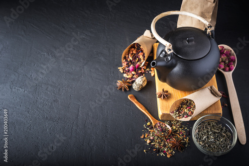 Black ceramic teapot with tea,Different tea and herbs , dried rose flowers on dark concrete background.Top view,copy space.