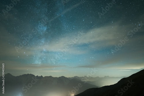 Milky Way galaxy and starry sky from high elevation in summertime on the Alps. Fog and mist, city lights.