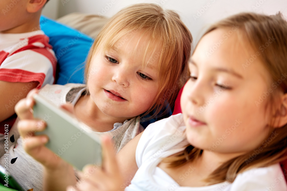 childhood, technology and family concept - happy little kids with smartphone in bed at home childhood, technology and family concept - happy little kids with smartphone in bed at home