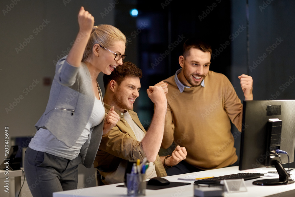 Naklejka premium business, success and teamwork concept - happy coworkers with computer celebrating victory at night office