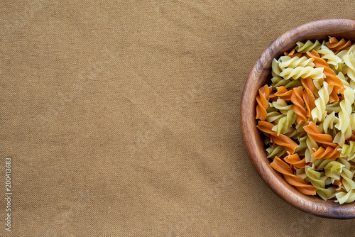 Multicolor spiral macaroni pasta in a wooden bowl on a brown rustic background texture with a side. Close-up with the top. With space for text.