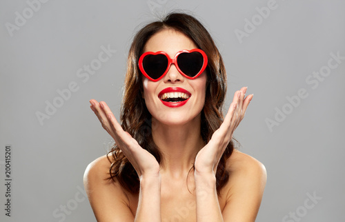 valentines day, beauty and people concept - happy smiling young woman with red lipstick and heart shaped sunglasses over gray background