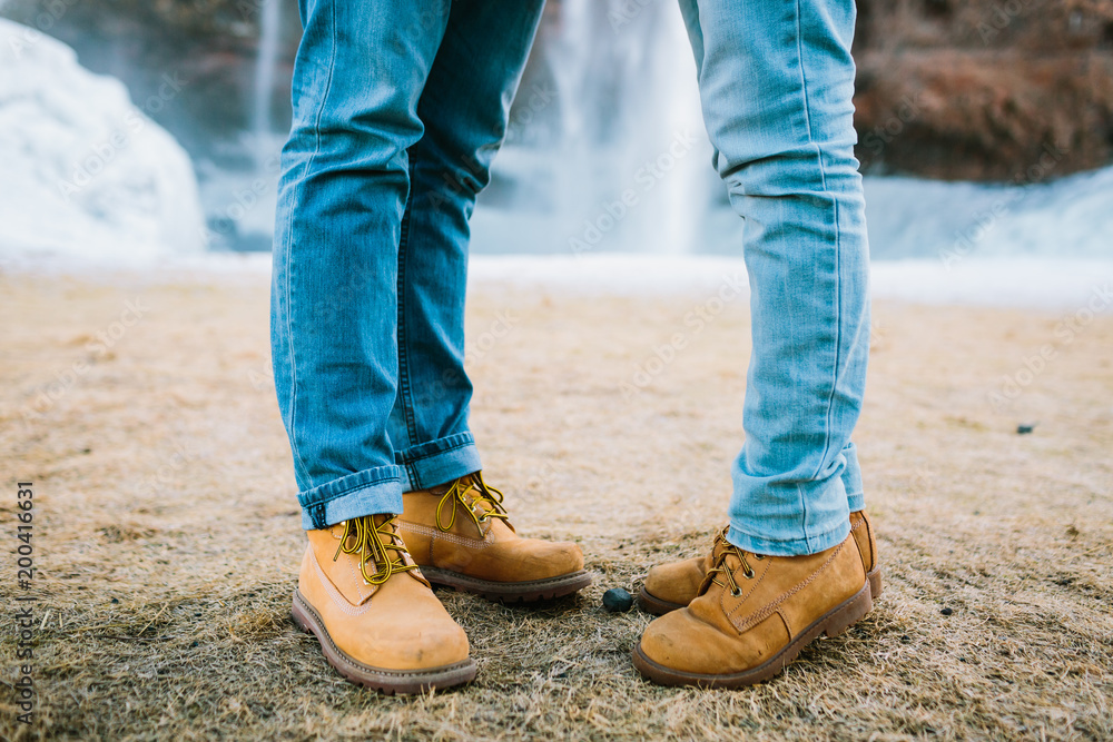 Couple in a brown hiking boots and blue jeans stand on waterfall background. Iceland. Copy space Couple in a brown hiking boots and blue jeans stand on waterfall background. Iceland. Copy space