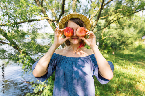 Happy woman in straw hat covering her eyes with peaches on garden background. Summer, outdoors. Close up portrait of young woman holding red peaches instead of her eyes. Organic food, fruit diet.