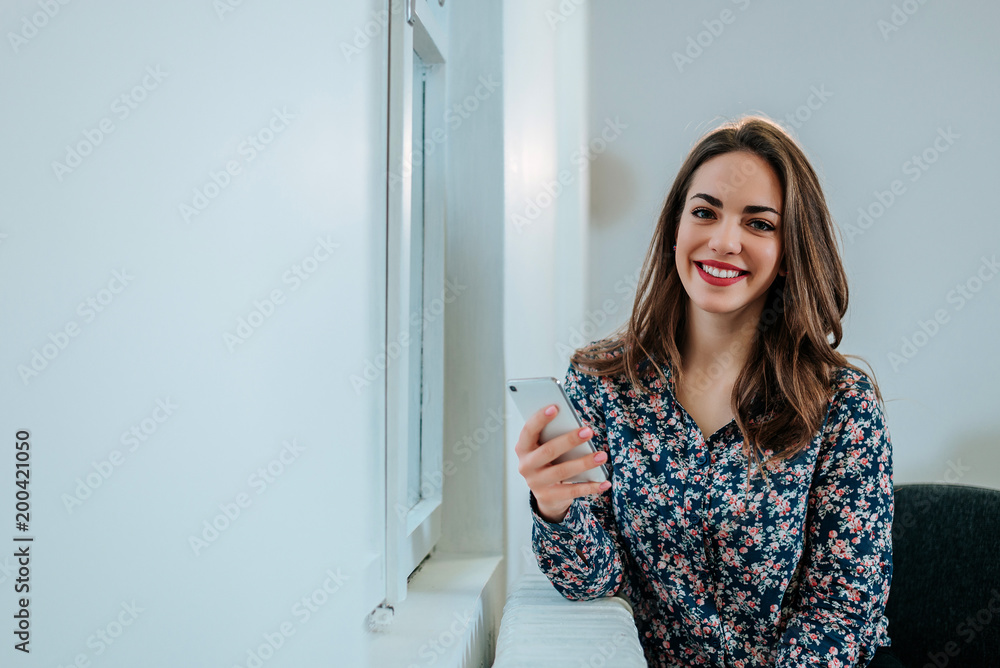 Attractive young woman with phone sitting near window.