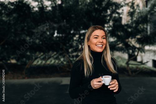 Portrait of a smiling blonde woman, in casual clothing, holding a cup of coffee while standing outdoors.