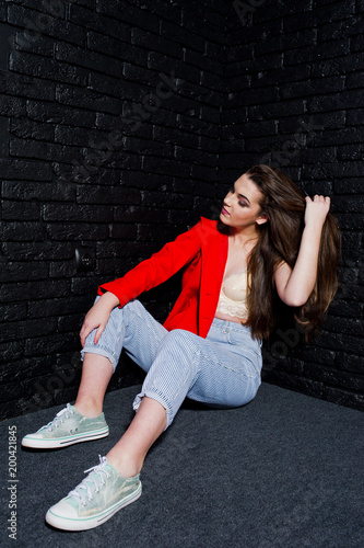 Stylish brunette girl on red jacket against studio black brick wall.