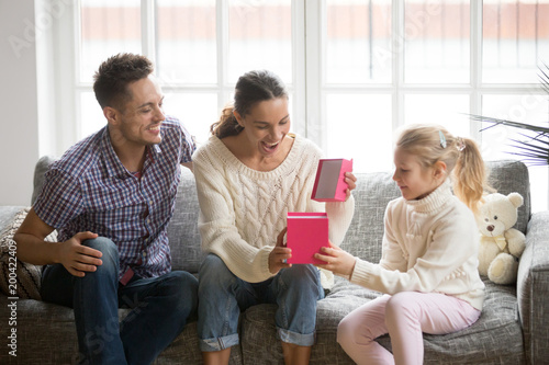 Excited woman opening gift box with present from child daughter and husband sitting on sofa together, kid girl congratulating happy young mom with birthday or mothers day making pleasant surprise