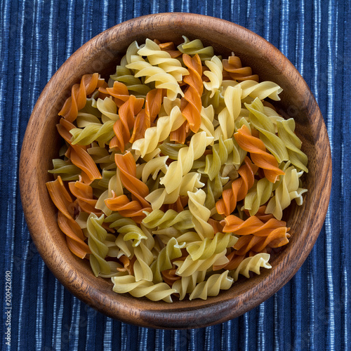 Multicolor spiral macaroni pasta in a wooden bowl on a striped white blue cloth background in the center. Close-up with the top.