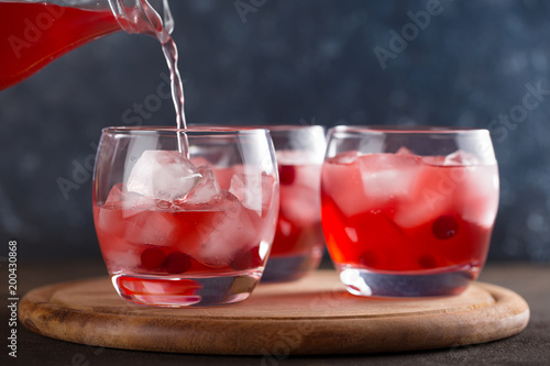 Cranberry juice pouring into glasses with berries and ice cube on the wooden cutting board.