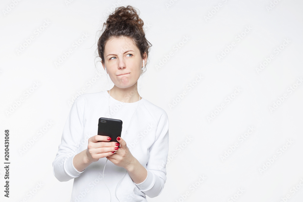 People, online communication and modern technologies concept. Thoughtful curly haired young hipster lady uses modern cell phone, listens to favorite music on earphones, uses free internet for player. People, online communication and modern technologies concept. Thoughtful curly haired young hipster lady uses modern cell phone, listens to favorite music on earphones, uses free internet for player.