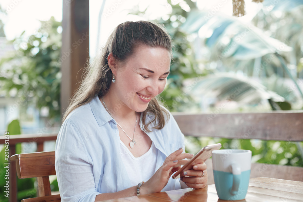 Cheerful brunette woman watches video in internet website, sends feed back on smart phone, dressed in casual clothing, drinks hot tea, recreates in outdoor cafeteria. People, leisure and technology.. Cheerful brunette woman watches video in internet website, sends feed back on smart phone, dressed in casual clothing, drinks hot tea, recreates in outdoor cafeteria. People, leisure and technology..