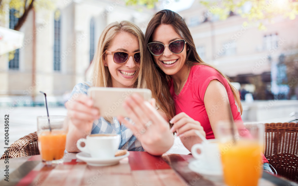 Friends meeting in a cafe. Young women drinking coffee and have fun with smart phone. Consumerism, lifestyle concept Friends meeting in a cafe. Young women drinking coffee and have fun with smart phone. Consumerism, lifestyle concept