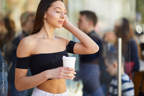 Brunette girl stand on the street look at camera and dring her delicious coffee from white cup, spring time photo shoot