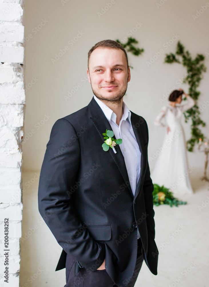 The bridegroom in a stylish suit stands in a white studio. Bride in the background.