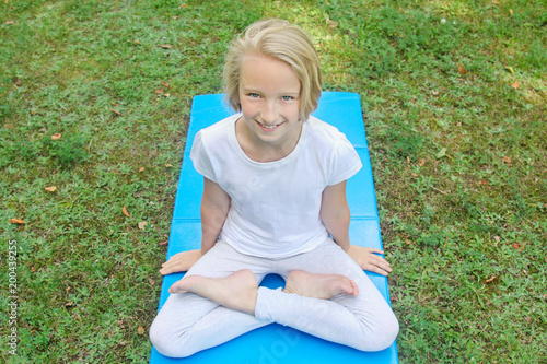 Beautiful blonde preteen girl in light clothing practicing yoga on a mat in the park. Healthy lifestyle. Outdoors workout. 