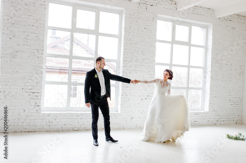 The bride and groom dancing by the big window in the studio with a white interior.