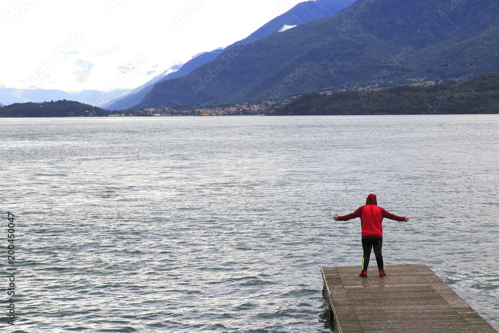 Mann steht auf einem Steg, Mit Jogginghose und Kaputzenshirt begleitet, am Comer See mit ausgebreiteten Armen und blickt nach Colico Mann steht auf einem Steg, Mit Jogginghose und Kaputzenshirt begleitet, am Comer See mit ausgebreiteten Armen und blickt nach Colico