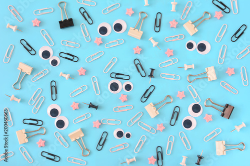 White and black paper clips, pink stars and stationery are scattered chaotically on a blue background. Funny smiley from clips in the center. Photo from the top.