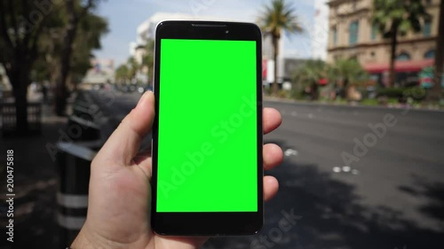 A man holds a smartphone outside near Las Vegas Boulevard as traffic and taxis pass by in the background. Green screen with optional corner pin markers for advanced screen replacement.  	