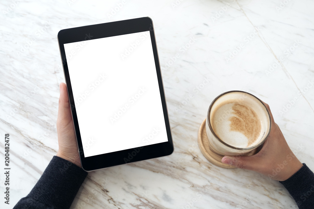 Top view mockup image of hands holding black tablet pc with white blank screen and coffee cup on table background Top view mockup image of hands holding black tablet pc with white blank screen and coffee cup on table background