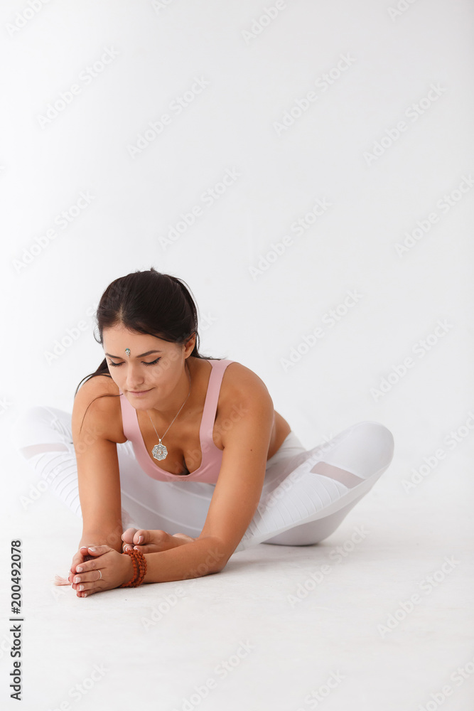 Portrait of young woman practicing yoga, sitting in Butterfly exercise, baddha konasana pose, working out, wearing white sportswear, indoor full length, home interior background Portrait of young woman practicing yoga, sitting in Butterfly exercise, baddha konasana pose, working out, wearing white sportswear, indoor full length, home interior background