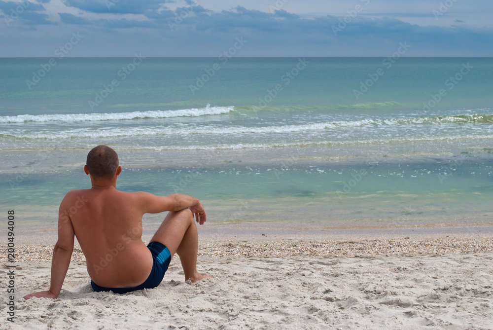young man sitting on the seashore from the back, summer beach waves sky with clouds, sand, sitting bending knee and looking at the horizon, resting on the summer weekend, vacation young man sitting on the seashore from the back, summer beach waves sky with clouds, sand, sitting bending knee and looking at the horizon, resting on the summer weekend, vacation