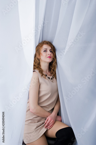 Curly hair girl on beige dress posed at white curtains.