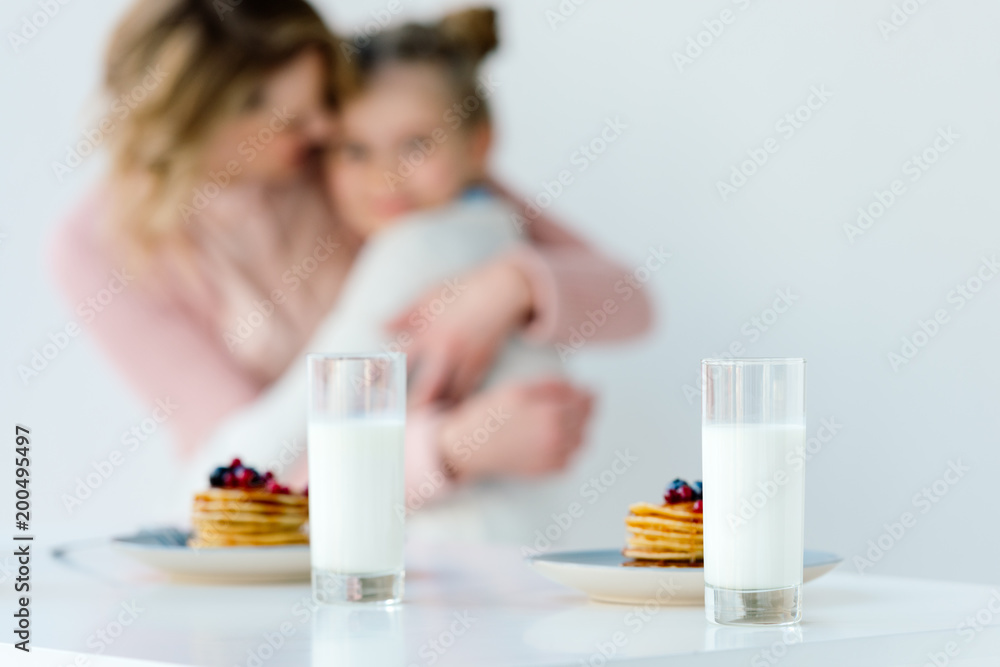 selective focus of glasses of milk and pancakes on tabletop with family hugging behind selective focus of glasses of milk and pancakes on tabletop with family hugging behind
