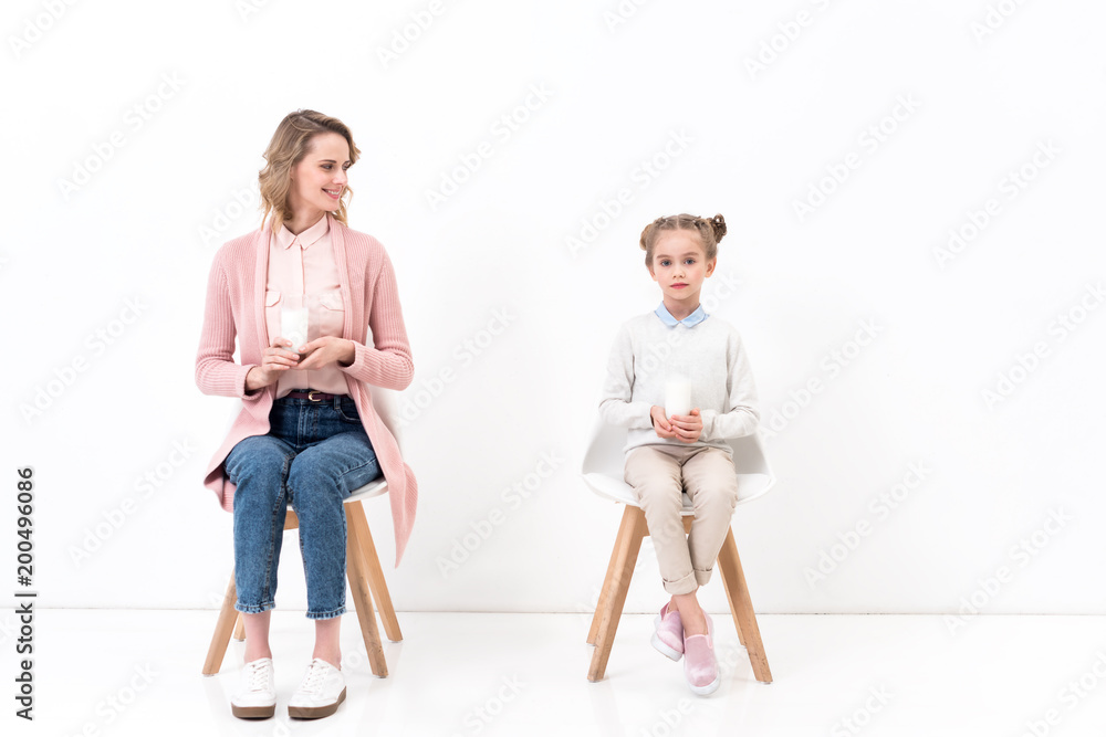 mother and daughter sitting on chairs with glasses of milk on white