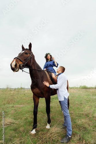 A happy girl is sitting on a horse and her boyfriend is standing by her in the middle of a meadow