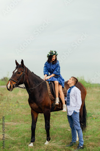 a pretty girl in a beautiful dresses elegantly sitting in a saddle on a horse and her boyfriend in an embroidered shirt stands next to her and they look at each other © Ivan