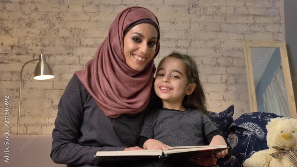 Young woman in hijab smiling and sits on sofa with her daughter and teaches her how to read, book, happy family concept, close up, home comfort in the background 50 fps Young woman in hijab smiling and sits on sofa with her daughter and teaches her how to read, book, happy family concept, close up, home comfort in the background 50 fps