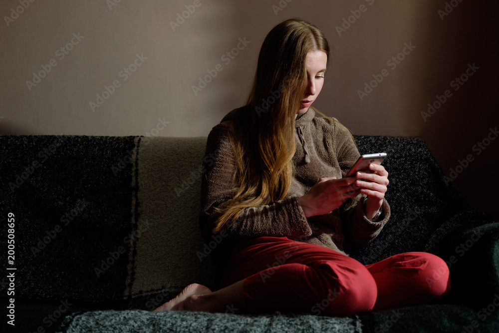 redhead ginger girl in hoodie reading message on smartphone, sitting on the sofa and smiling redhead ginger girl in hoodie reading message on smartphone, sitting on the sofa and smiling