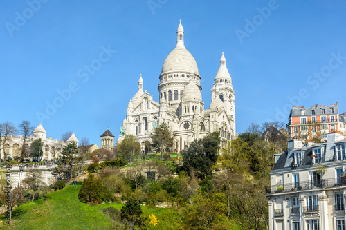View of the basilica of the Sacred Heart of Paris on top of the Montmartre hill under clear blue skies with the Louise Michel park below and apartment buildings in the foreground.