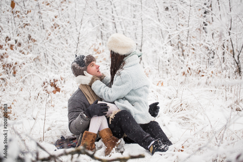 The happy young man sitting in the snow-covered park and holding the young woman on the lap and they look at each other The happy young man sitting in the snow-covered park and holding the young woman on the lap and they look at each other