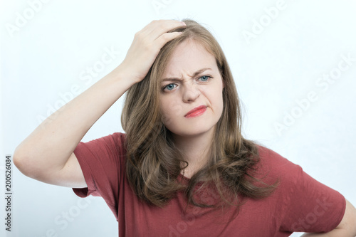 Portrait of blonde girl or young woman wearing red t-shirt with perplexed, puzzled, dumb, ignorant expression on her face scratching head with hand. When you don't know the answer