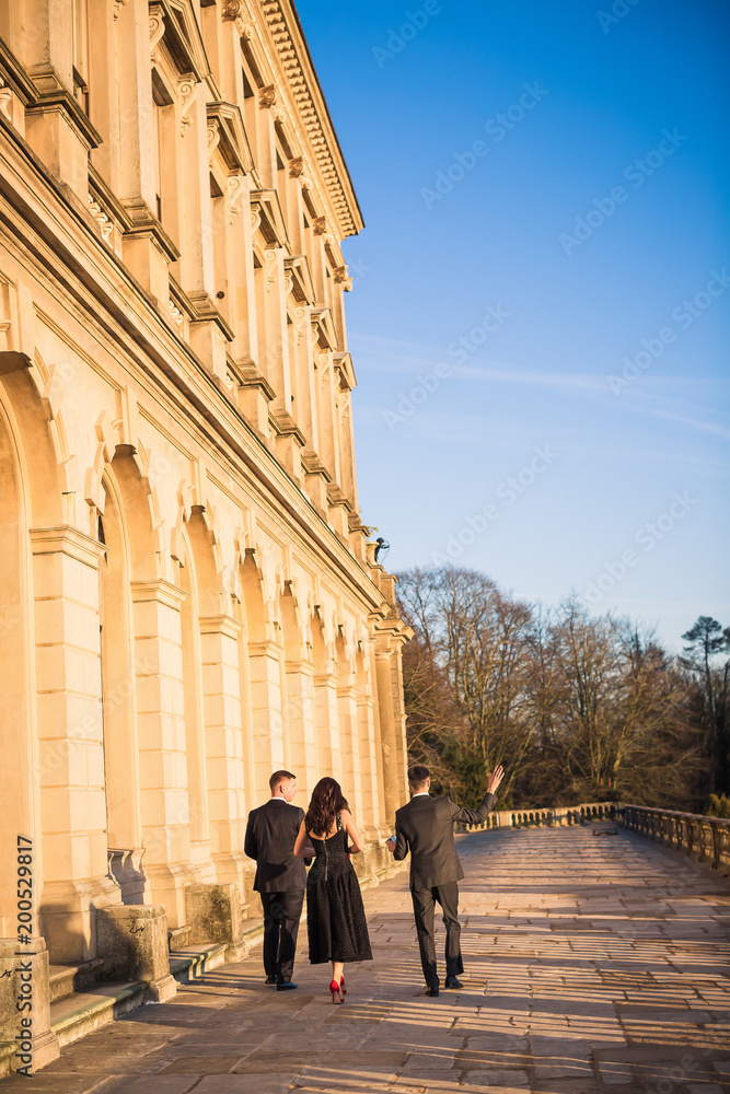 Back view of the young couple and young man near them who are walking together by the palace