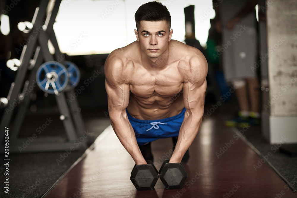 Fototapeta premium Strong, handsome man doing push-ups on dumbbells in a gym as bodybuilding exercise, training his muscles