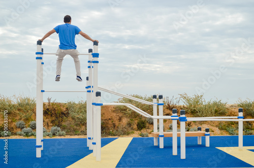 Outdoors fitness training active lifestyle, back view of man doing sport exercising on horizontal bar in summer park sky background